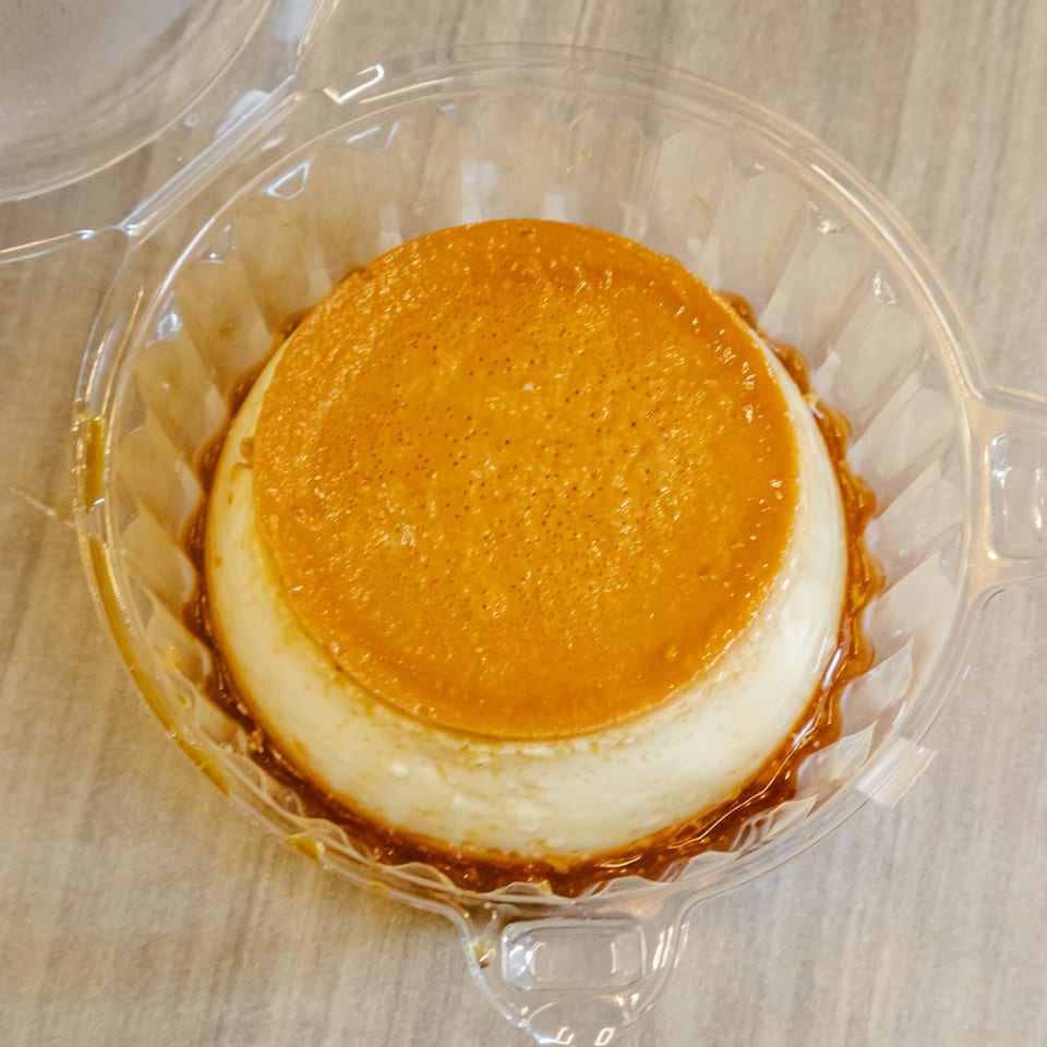Caramel-topped cheesecake in a clear plastic container on a beige surface.