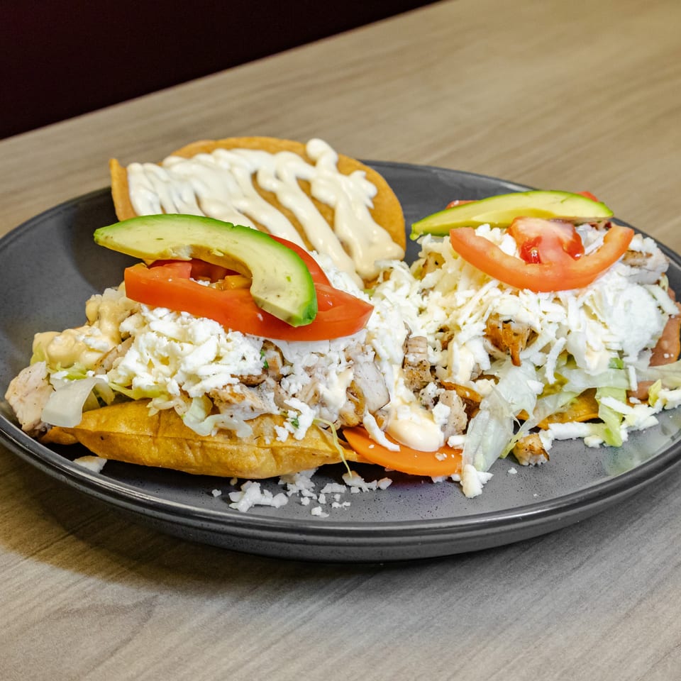 Loaded tostadas topped with grilled chicken, lettuce, tomatoes, avocado, crumbled cheese, and creamy sauce on a black plate.
