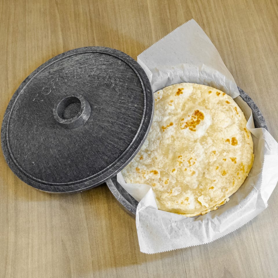 Round flatbread with toasted spots served in a paper-lined black basket beside its lid on a wooden table.
